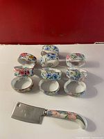 Photo shows 10 porcelain napkin rings arranged in two rows, each decorated with colorful floral designs and gold trim, along with one stainless steel cheese cutter with a floral handle.