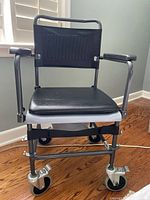 Front view of a black and gray rolling commode chair with padded seat and backrest, plastic armrests, and locking casters