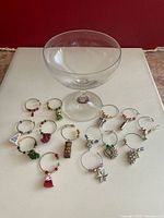 Photo showing one clear stemmed glass and 15 assorted metal wire wine glass charms with decorative glass beads and charms laid out on a white surface