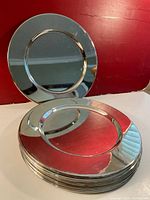 Photo of eight silver-colored metal charger plates stacked with one plate standing upright behind them. Plates have a reflective smooth finish.