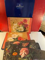 Set of placemats partially inside one original blue Pimpernel box, showing light floral designs with roses and white flowers.