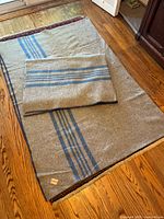 Photo showing two tan/grey wool blankets with blue stripes laid out on a wooden floor, showcasing size and pattern.