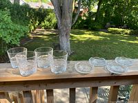 Four clear crystal glasses and four clear crystal coasters arranged in a line outdoors on a wooden rail with green garden background.