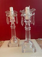 Front view of two clear crystal Victorian-style candlestick holders with ruffled tops and hanging prisms, each holding a partially used white candle.