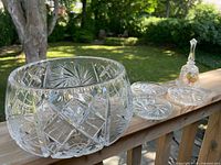 Wide shot of the large crystal bowl, three matching coasters with pinwheel patterns, and a clear Cuba Bell with fruit designs set outdoors on wooden railing.