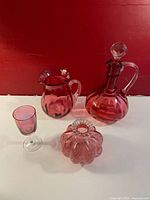 Photo showing all five pieces: decanter with stopper, jug, small glass, and a decorative vase; set on a white surface with a red background.