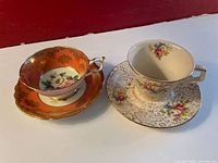 Two vintage bone china teacup and saucer sets shown from the side, one white with floral designs and the other orange with gold accents and floral motif inside the teacup.