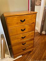 Front view of wooden dresser showing 5 drawers with black ornate handles and light to medium wood finish.