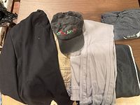 All five items folded: black blazer, khaki pants, light grey pants, dark grey Laredo T-shirt, black "Believe" cap