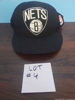 Front view of black Brooklyn Nets cap with white embroidered logo and NBA patch on side, shown on wooden surface with lot number card.