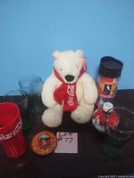 Full view of the lot: white Coca-Cola plush bear, multiple Coca-Cola branded glasses, a travel mug, a shaker bottle, and a red tumbler arranged on a flat surface against a blue backdrop.