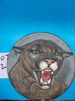 Front view of round ceramic plate with hand painted cougar face; mouth open showing teeth, pink nose, yellow eyes.