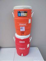 Photo showing two stacked large sports water jugs, one bright orange on top and one red on bottom, both with white lids and handles, no visible damage