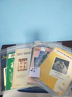 Photo of all 10 vintage sheet music pieces stacked in plastic sleeves showing varied colorful covers.