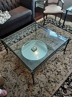 Full view of the square glass top coffee table placed on a patterned rug with couch and chairs around it.