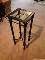 Front angled view of black metal plant stand with clear square glass top showing leg design and frame.