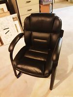 Single black leather chair with padded arms and backrest, metal legs shown on carpeted floor with framed items behind.