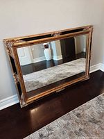 Mirror leaning against wall horizontally showing ornate black and gold frame details and clear reflective surface.
