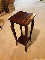 Single wooden plant stand with curved legs, square top, and lower shelf on a light carpet.