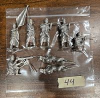 Eight vintage metal miniatures unpainted, displayed in a clear plastic bag on a wooden surface. Includes various soldier poses with weapons and a flag.