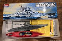 Open box of Academy German Battleship Bismarck plastic model kit showing assembled hull with motor and battery, sprues with parts, and instruction sheet inside the box under the hull