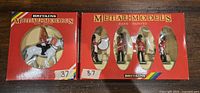 Photo showing two unopened original boxes of Britains Metal Models with hand-painted miniatures visible through display windows. One box contains a mounted soldier on white horse, the other contains three standing soldiers including one with a drum.