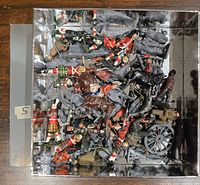 Top-down photo of a box full of Britains lead military miniature figures including painted and unpainted soldiers, horses, and a cannon.