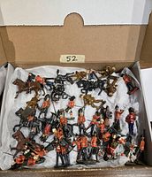 Box displaying painted lead miniature military figures including British Royal Guard infantry and cavalry, some horse-mounted, arranged on tissue paper inside a cardboard box.
