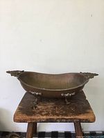 Front-facing view of antique metal basin placed on a wooden stool, showing detailed feet and side handle.