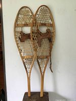 Pair of antique wooden frame snowshoes with woven natural fiber netting and leather foot bindings hanging on a white wall.
