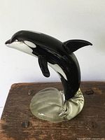 Solid art glass killer whale statue, side view showing black and white orca leaping from clear glass base on wooden surface.