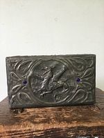 Front view of dark metal rectangular box with raised dragon figure encircled by carved patterns and two blue jewelled accents on the front face.