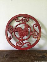 Front view of round cast iron trivet with central rooster design and decorative scrolls, painted red.