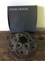 Top view of the circular dark metal ornament showing nine rounded protrusions and teardrop cut-out holes, placed in front of the Dansk Designs branded box.
