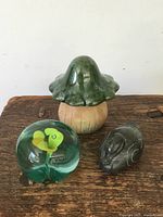 Photo showing the three desk ornaments: green glass paperweight with internal yellow flowers, dark stone duck figurine, and green-topped pottery mushroom with a brown stem on a wooden surface.