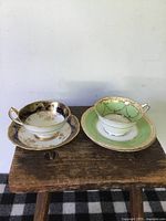 Two porcelain tea cups and saucers on a wooden surface, one in dark blue with elaborate gold floral designs and one in green with gold chain-like accents.