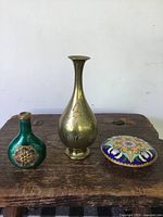 Three antique Asian decorative metal items displayed together on wooden surface: green enamel snuff bottle missing lid, brass bud vase, and cloisonné lidded box.
