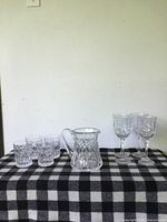 Wide shot of all crystal pieces on a black and white checkered cloth including four short tumblers, a pitcher, and two wine glasses