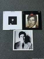 Three Leonard Cohen vinyl records with original covers laid out on carpet showing front side with portraits and art.
