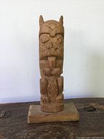 Front view of small carved wooden totem pole showing detailed facial and body features, mounted on a rectangular base.