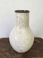 Front view of cream glazed vase with raised floral detail and brown glazed rim seen against white background on rustic wood surface.
