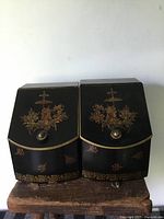 Two black antique tin canisters side by side, front view showing painted Asian theme designs, metal knobs and decorative trim