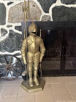 Front view of the vintage brass knight suit of armor holding a polearm, standing on a hexagonal base in front of a fireplace.