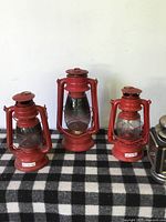 Frontal photo showing three red vintage metal and glass lanterns of varying sizes arranged on a black and white checked cloth.