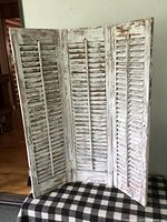 Three-panel white painted rustic folding shutter standing open on a checkered blanket, showing distressed and peeling paint surface