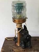 Front-angle view of vintage Scotty dog lamp with black dog figurine base, brass cylindrical support, textured glass lampshade, and coiled electrical cord.