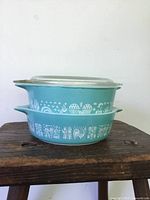Side view of two turquoise Pyrex canisters stacked, showing white folk art decorative patterns and glass lids.