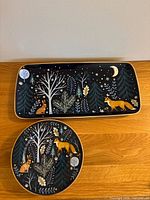 Rectangular and round ceramic plates with forest animal and nature design, gold rims, placed on wooden surface.