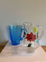 Photo showing both blue hobnail glass tumbler and clear hand-painted floral pitcher side-by-side on a white surface with neutral background.