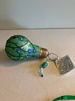 Side view of the decorative hand-painted glass light bulb showing painted blue flowers, green vines, a white fairy, and a ladybug with a gold-colored metal screw base and attached beaded ornament.